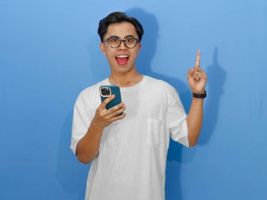 Beyaz tişörtlü heyecanlı genç Asyalı adam cep telefonuyla poz veriyor, sanki aklına harika bir fikir gelmiş gibi..