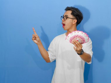 Beyaz tişörtlü heyecanlı Asyalı genç adam, neşeyle elinde para tutuyor ve bir kenara, finansal temalar için mükemmel..