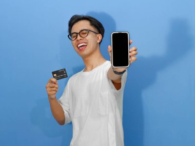 Beyaz gömlekli heyecanlı genç Asyalı adam elinde bir kredi kartı tutarken boş telefon ekranı gösteriyor. Reklamlar ve promosyonlar için mükemmel..