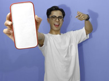 Beyaz tişörtlü heyecanlı Asyalı genç adam boş bir cep telefonu ekranını işaret ediyor. Promosyon, reklam veya dijital pazarlama içeriği için mükemmel..