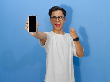 Beyaz tişört giyen genç bir Asyalı 'nın portresi. Başarı kutlamalarında yumruklarını sıkarken boş bir cep telefonu ekranını heyecanla gösteriyor..