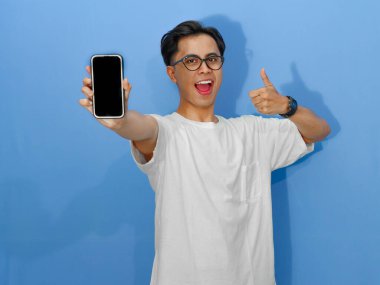 Beyaz tişörtlü heyecanlı genç Asyalı adam boş bir cep telefonu ekranını gösterirken başparmak kaldırıyor, reklam ve promosyonlar için mükemmel..