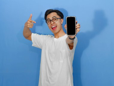 Beyaz tişörtlü heyecanlı Asyalı genç adam boş bir cep telefonu ekranını işaret ediyor. Promosyon, reklam veya dijital pazarlama içeriği için mükemmel..