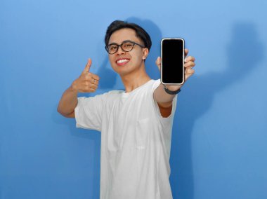 Beyaz tişörtlü heyecanlı genç Asyalı adam boş bir cep telefonu ekranını gösterirken başparmak kaldırıyor, reklam ve promosyonlar için mükemmel..