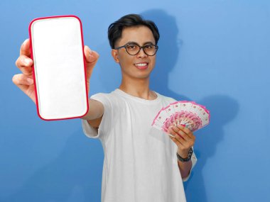 Asyalı genç bir adam mutlu, heyecanlı bir yüz ifadesiyle elinde para tutarken boş bir cep telefonu ekranı gösteriyor, mavi bir arka planda izole edilmiş. Reklamlar için mükemmel..