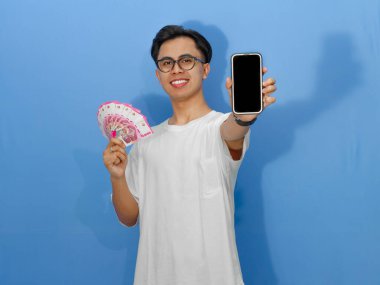 Asyalı genç bir adam mutlu, heyecanlı bir yüz ifadesiyle elinde para tutarken boş bir cep telefonu ekranı gösteriyor, mavi bir arka planda izole edilmiş. Reklamlar için mükemmel..