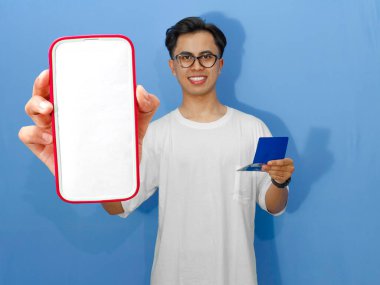 Beyaz gömlekli genç Asyalı adam elinde banka tasarruf defteri tutarken boş bir cep telefonu ekranı gösteriyor. Reklam, finans veya bankacılık kavramları için mükemmel..