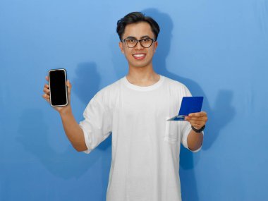 Beyaz gömlekli genç Asyalı adam elinde banka tasarruf defteri tutarken boş bir cep telefonu ekranı gösteriyor. Reklam, finans veya bankacılık kavramları için mükemmel..