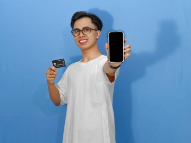 Beyaz gömlekli heyecanlı genç Asyalı adam elinde bir kredi kartı tutarken boş telefon ekranı gösteriyor. Reklamlar ve promosyonlar için mükemmel..