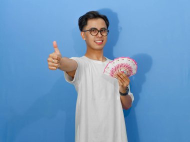 Beyaz tişörtlü mutlu bir Asyalı adam para gösteriyor ve başarıyı ve neşeyi simgeliyor..