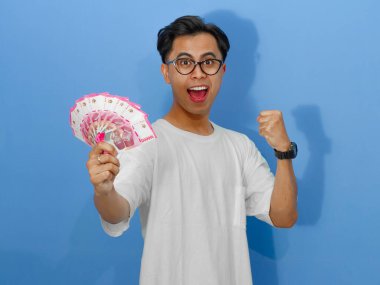 Beyaz tişörtlü heyecanlı Asyalı genç adam para tutarken başarıyı kutluyor, neşe ve başarı saçıyor..