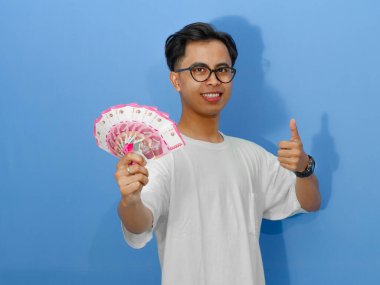 Beyaz tişörtlü mutlu bir Asyalı adam para gösteriyor ve başarıyı ve neşeyi simgeliyor..