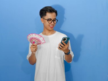 Beyaz tişörtlü genç Asyalı bir adamın portresi, cep telefonu ve para tutarken gülümsüyor, güven ve pozitiflik yayıyor..