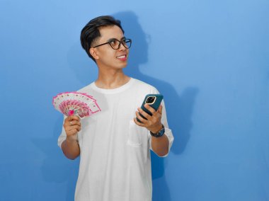 Beyaz tişörtlü genç bir Asyalı elinde para ve cep telefonu tutarken gülümsüyor, sola bakıyor. Finans ve yaşam tarzı temaları için mükemmel.