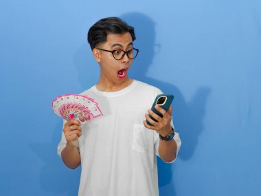Beyaz tişörtlü genç Asyalı bir adam elinde para tutarken ve akıllı telefonuna bakarken komik bir sürpriz ifade sergiliyor..