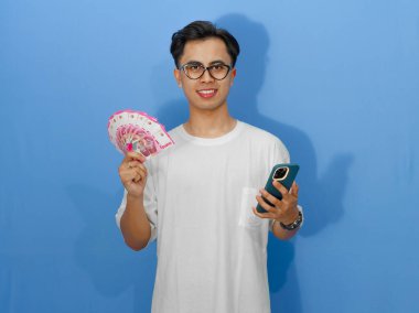Beyaz tişörtlü mutlu bir Asyalı adam, elinde cep telefonu ve para tutarken gülümsüyor, yaşam tarzı ve finansal temalar için mükemmel..
