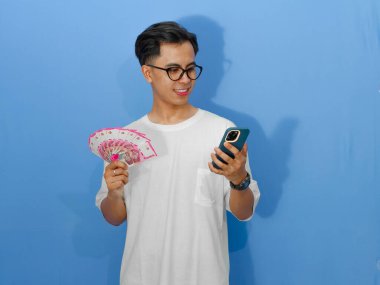 Beyaz tişörtlü genç Asyalı bir adamın portresi, cep telefonu ve para tutarken gülümsüyor, güven ve pozitiflik yayıyor..