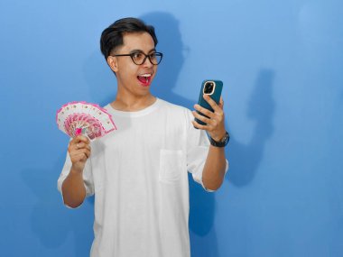 Beyaz tişörtlü genç Asyalı bir adam elinde para tutarken ve akıllı telefonuna bakarken komik bir sürpriz ifade sergiliyor..