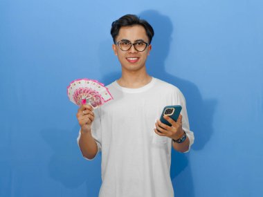 Beyaz tişörtlü mutlu bir Asyalı adam, elinde cep telefonu ve para tutarken gülümsüyor, yaşam tarzı ve finansal temalar için mükemmel..