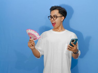 Beyaz tişörtlü genç Asyalı bir adam elinde para tutarken ve akıllı telefonuna bakarken komik bir sürpriz ifade sergiliyor..