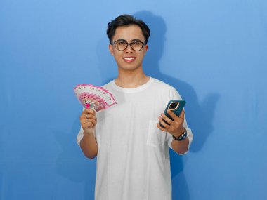 Beyaz tişörtlü mutlu bir Asyalı adam, elinde cep telefonu ve para tutarken gülümsüyor, yaşam tarzı ve finansal temalar için mükemmel..