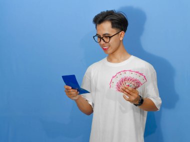 Banka tasarruf defteri ve para tutarken gülümseyen Asyalı genç bir adamın portresi mutluluğu ifade ediyor. Finansal temalar ve yaşam tarzı kavramları için ideal.