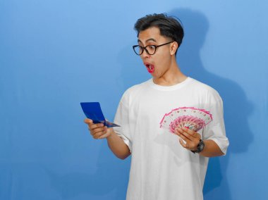 Beyaz tişörtlü genç bir Asyalı elinde banka tasarruf defteri ve para tutarken çok komik bir ifade sergiliyor.