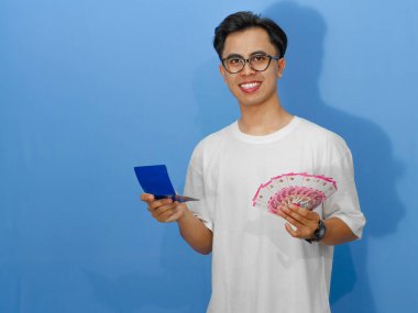 Banka tasarruf defteri ve para tutarken gülümseyen Asyalı genç bir adamın portresi mutluluğu ifade ediyor. Finansal temalar ve yaşam tarzı kavramları için ideal.