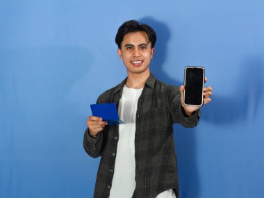 Confident young Asian man in casual wear holds bank saving book and shows blank smartphone screen, ideal for financial and tech concepts. clipart