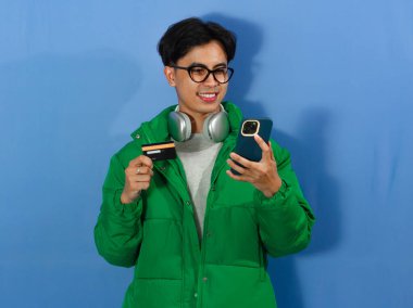 Yeşil kışlık ceketli gülümseyen Asyalı genç adam, akıllı telefon kullanıyor ve elinde bir kredi kartı tutuyor..