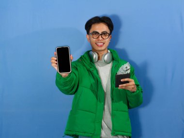 Kışlık ceketli genç Asyalı adam boş telefon ekranı ve içi para dolu cüzdan taşıyor. Neşeli bir gülümseme, reklam ve terfi için ideal..