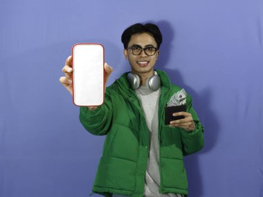 Kışlık ceketli genç Asyalı adam boş telefon ekranı ve içi para dolu cüzdan taşıyor. Neşeli bir gülümseme, reklam ve terfi için ideal..