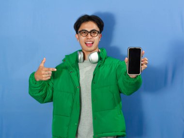 Kışlık ceketli genç Asyalı adam heyecanlı bir ifadeyle boş telefon ekranını işaret ediyor. Reklam, promosyon ve dijital pazarlama için ideal..