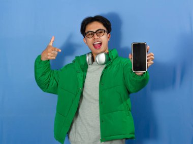 Kışlık ceketli genç Asyalı adam heyecanlı bir ifadeyle boş telefon ekranını işaret ediyor. Reklam, promosyon ve dijital pazarlama için ideal..