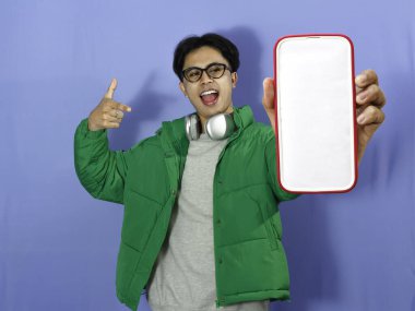 Young Asian man in winter jacket pointing at blank phone screen with excited expression. Ideal for advertising, promotions, and digital marketing. clipart