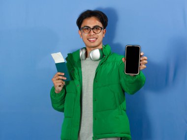 Elinde pasaport tutarken gülümseyen ve seyahat temaları için ideal olan boş bir cep telefonu ekranı gösteren kışlık ceketli Asyalı bir adamın portresi..