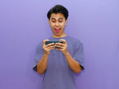 Heyecanlı genç Asyalı adam telefonuyla mobil oyunlar oynuyor, canlı mor bir arka plana karşı neşe ve nişan sergiliyor..