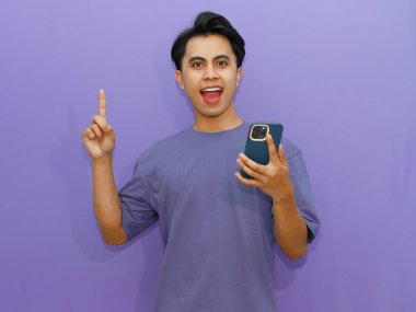 Heyecanlı genç Asyalı adam canlı mor bir arka plana karşı harika bir fikri kutlamak için cep telefonuyla işaret ediyor..