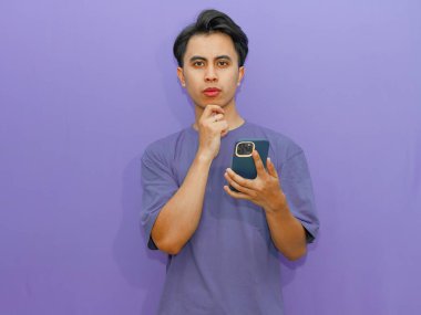 Elinde düşünceli bir ifadeyle cep telefonu tutan genç bir Asyalı. Gözleri derin derin bakıyor. Mor bir arka planda izole edilmiş..