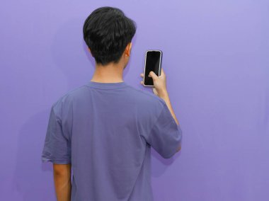 Genç Asyalı bir adamın cep telefonunu canlı bir mor arkaplana karşı kullanmasının arkasında modern yaşam tarzı ve teknolojisi sergileniyor..