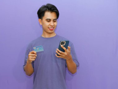 Genç Asyalı adam kendine güvenen bir kimlik kartı ve cep telefonu tutuyor, canlı mor bir arka plana gülümsüyor..