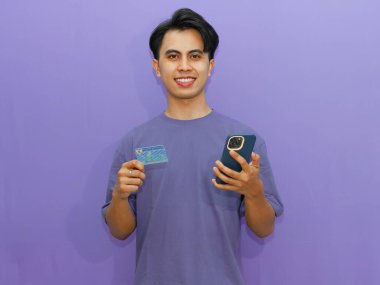 Genç Asyalı adam kendine güvenen bir kimlik kartı ve cep telefonu tutuyor, canlı mor bir arka plana gülümsüyor..