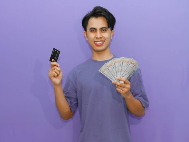 Asyalı genç bir adam elinde nakit para ve kredi kartı tutuyor, kendine güveni tam, mor arka planda izole edilmiş, başarı yayıyor..