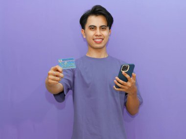 Genç Asyalı adam güven içinde kimlik kartını ve cep telefonunu gösteriyor. Mor bir arka planda gülümsemesi var..