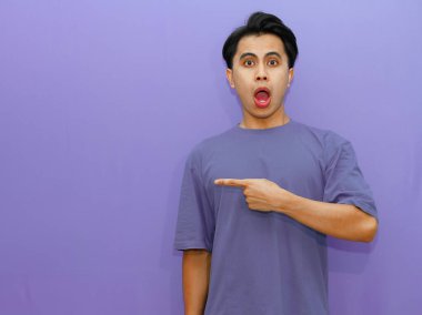Şok olmuş Asyalı bir adam canlı mor bir arka plana karşı yan tarafı işaret ederek sürpriz ve mizah anı yakalıyor..