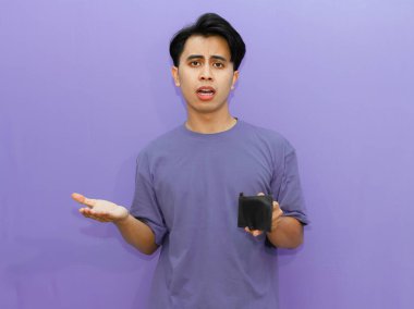Zavallı Asyalı genç bir adam, boş bir cüzdanı var, üzgün ve şok olmuş bir ifade sergiliyor, izole bir mor arka plana karşı..