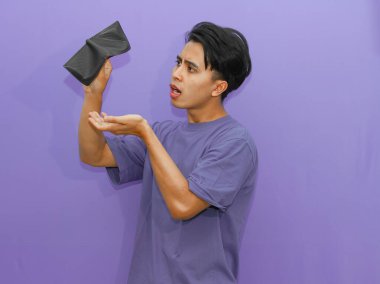 Cüzdanı boş üzgün bir Asyalı genç adam. Mor arka planda stres gösteriyor. Mali sıkıntıları ve zorlukları gösteriyor..