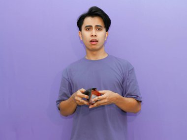 Poor young Asian man with an empty wallet, displaying a sad and shocked expression against an isolated purple background. clipart