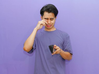 Zavallı Asyalı genç adam boş bir cüzdan tutuyor, mor arka planda ağlayan bir jest gösteriyor, finansal zorlukları vurguluyor..