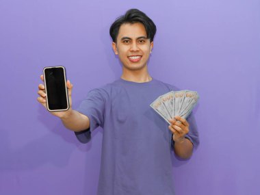 Gülümseyen Asyalı genç bir adam, canlı mor bir arkaplan üzerine kurulmuş banknotları tutarken boş bir cep telefonu ekranı gösteriyor..
